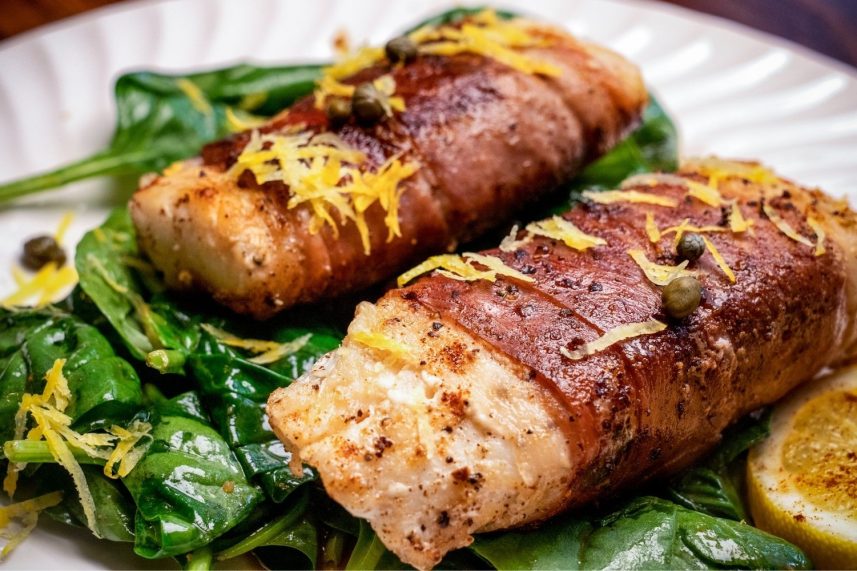 Plato con dos peces envueltos en tocino, rodeados de espinacas verdes.